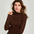 A woman wearing a ribbed brown mock neck long-sleeve top, holding the collar slightly with her hand against a plain light gray background.