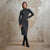 A woman stands against a textured beige plaster wall wearing a fitted dark gray long-sleeve midi dress with a thin black belt, paired with black knee-high boots.