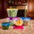 A set of clear plastic food storage containers with colorful lids in green, purple, blue, and pink, arranged on a patterned beige table in front of a dark wood cabinet and fireplace.