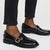A person wearing black leather Mary Jane loafers with gold-tone buckle straps and dark wash cropped jeans, shown from the calves down on a light gray studio background.