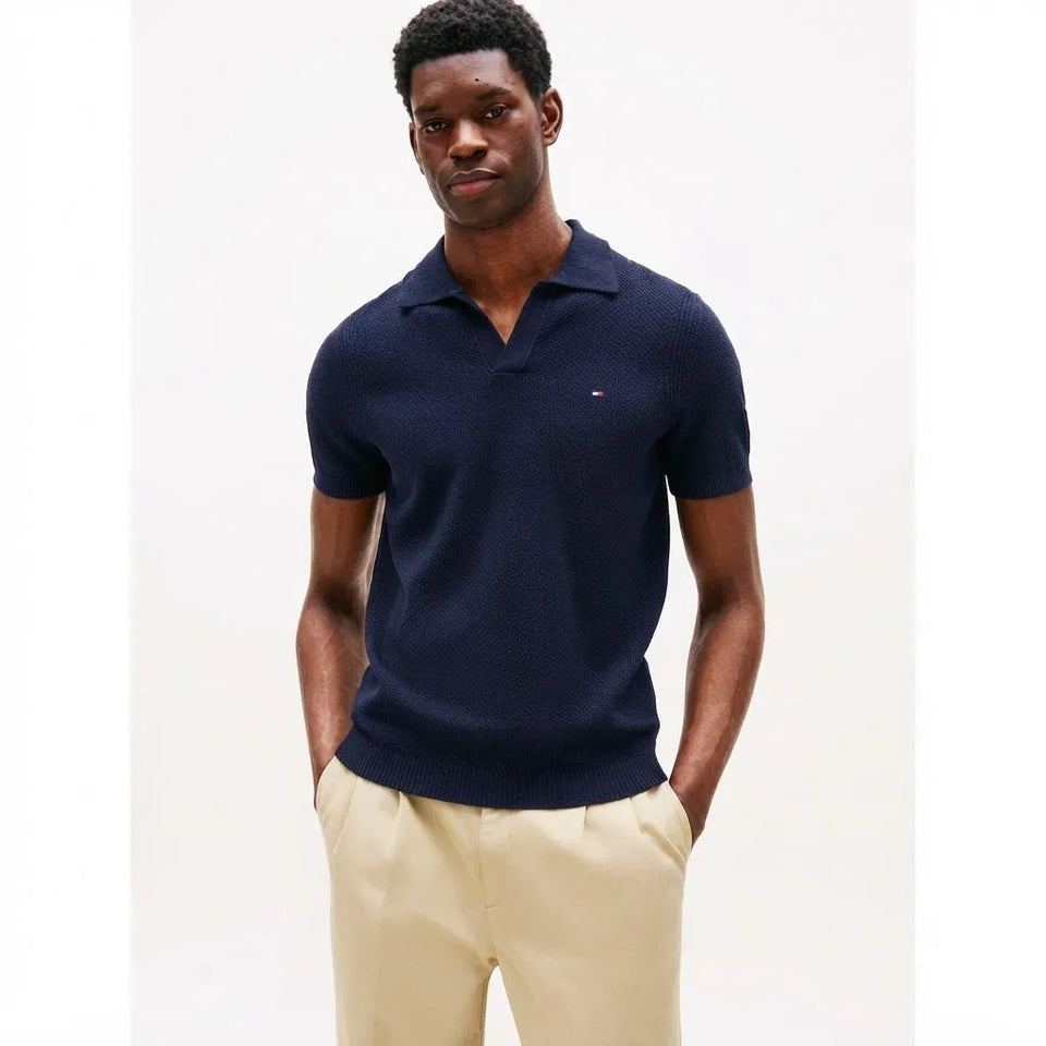 A man wearing a navy blue short-sleeve knit polo shirt with a small embroidered Tommy Hilfiger flag logo on the chest, paired with beige trousers, against a plain white studio background.