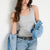 A woman wearing a light gray ribbed tank top, light-wash blue jeans, a black studded belt, and holding a light blue denim shirt over her shoulder, posed against a plain white wall.