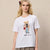 A blonde woman modeling a white short-sleeve graphic T-shirt with a teddy bear illustration and the text 'Ought at the week' on the front, paired with a pleated brown skirt against a textured beige wall.