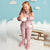 A young girl stands in a studio setting wearing a shiny metallic pink puffer snowsuit with matching pink boots, holding a white ball. The background features a light blue sky backdrop with white cloud cutouts, a wooden bench, and a white textured floor.