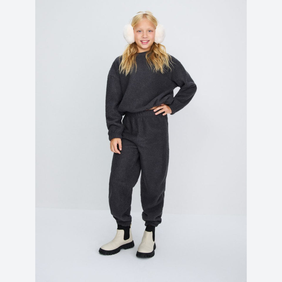 A young girl modeling a dark gray heathered two-piece sweatshirt and jogger pants set, styled with white earmuffs and off-white boots, against a light gray studio background.