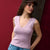 A woman wearing a light purple sleeveless V-neck tank top and blue jeans, posing against a dark red and black background.