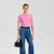 A woman modeling a short-sleeve pink crewneck t-shirt, paired with high-waisted blue jeans, a black belt, and holding a small black handbag against a light gray studio background.