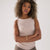 A woman with curly brown hair wearing a ribbed sleeveless top in a light beige color and brown pants, standing against a plain light gray background.