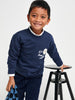 Child wearing a navy sweater with a logo, standing next to a black stool on a white background