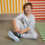 A young boy with curly brown hair sits on a gray concrete floor, wearing a light gray sweatshirt and matching sweatpants, while tying a black and neon yellow sneaker. The background features a yellow and white striped wall on the left and a red and white striped wall on the right, with a green and white striped rug on the floor.
