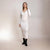 A woman modeling a white ribbed knit long-sleeve midi dress with a wrap-style V-neck bodice, paired with black ankle boots, against a plain light beige studio backdrop.