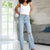 A woman modeling light-wash blue high-rise flare jeans with distressed knee details, paired with a white tank top and black sandals, standing in a modern living room setting.