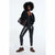 A model with curly black hair stands against a white studio background wearing shiny black leggings, a black bomber jacket, brown suede boots, white socks, and carrying a dark brown leather tote bag.