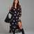A woman models a black long-sleeve midi dress with a pink floral print, styled with a large black textured shoulder bag and black knee-high boots against a solid gray studio background.