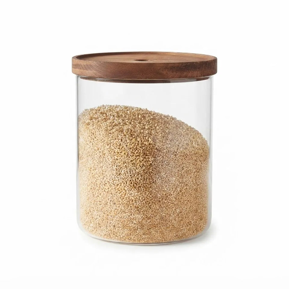 A clear glass storage jar with a round wooden lid, filled with light tan grains or seeds, shown on a plain white background.