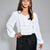 A woman modeling a white long-sleeve blouse with a smocked waist and ruffled hem, paired with a black skirt, against a neutral gray studio background.