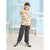 A child model wearing a cream-colored t-shirt with a colorful cartoon character print, dark gray jogger pants, and black-and-white sneakers, posing in front of a light blue and white shelving background.