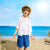 A young boy stands on a sandy beach wearing a straw hat, a white short-sleeve button-up shirt over a white t-shirt, and blue cargo shorts, with the ocean and blue sky behind him.