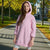 A young girl wearing a light pink oversized hooded sweatshirt with rolled-up sleeves and a front kangaroo pocket, standing on a paved path in an outdoor park setting with autumn trees.