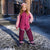 A young girl stands outdoors on a snowy city street wearing a pink and magenta winter snowsuit with a matching pink beanie and dark snow boots.