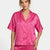 A woman wearing a bright pink short-sleeve satin pajama top with an allover tonal heart pattern, paired with matching pink pajama pants, against a plain light gray background.