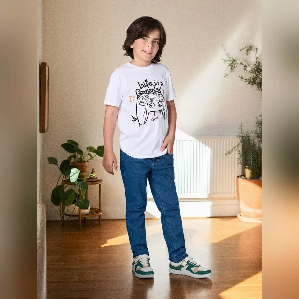 A child wearing a white graphic t-shirt with Arabic text, blue jeans, and green and white sneakers, standing in a sunlit room with plants and a radiator.