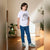 A child wearing a white graphic t-shirt with Arabic text, blue jeans, and green and white sneakers, standing in a sunlit room with plants and a radiator.