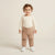 A toddler standing against a warm beige studio background, wearing a cream long-sleeve top and tan jogger pants with embroidered animal faces near the lower legs, paired with white sneakers.