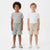 Two children standing side by side wearing matching short-sleeve t-shirts and drawstring shorts. The child on the left wears a heather gray t-shirt with sage green shorts, and the child on the right wears a white t-shirt with beige shorts.