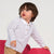 A toddler boy wearing a white long-sleeve polo shirt with thin dark stripes and tan pants, leaning on a bright pink sculptural chair against a light gray background.