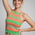 A smiling woman wearing a ribbed knit halter crop top with horizontal stripes in green, orange, and pink, paired with light beige pants, against a solid light gray background.