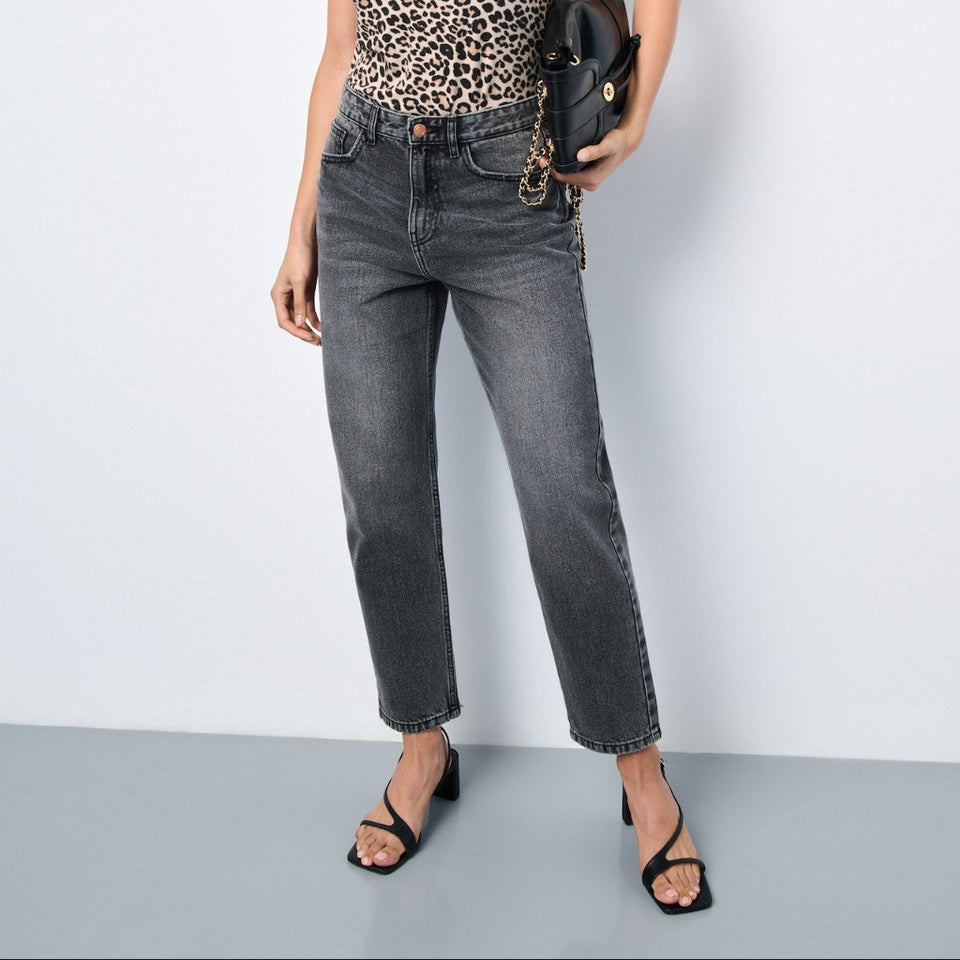 A model stands against a light gray studio wall wearing a leopard-print spaghetti-strap camisole top, dark gray cropped jeans, black strappy heels, and holding a black leather clutch with gold hardware.