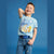 A young boy smiling and posing in a light blue graphic t-shirt and blue jeans against a solid blue studio background.
