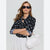 A woman wearing a black long-sleeve blouse with white polka dots and a keyhole cutout, styled with a white skirt, black sunglasses, and a white shoulder bag against a light gray studio background.