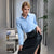 A woman posing in a light blue long-sleeve button-up shirt and a high-waisted black faux-leather skirt with a thigh slit, set in a modern living room environment.