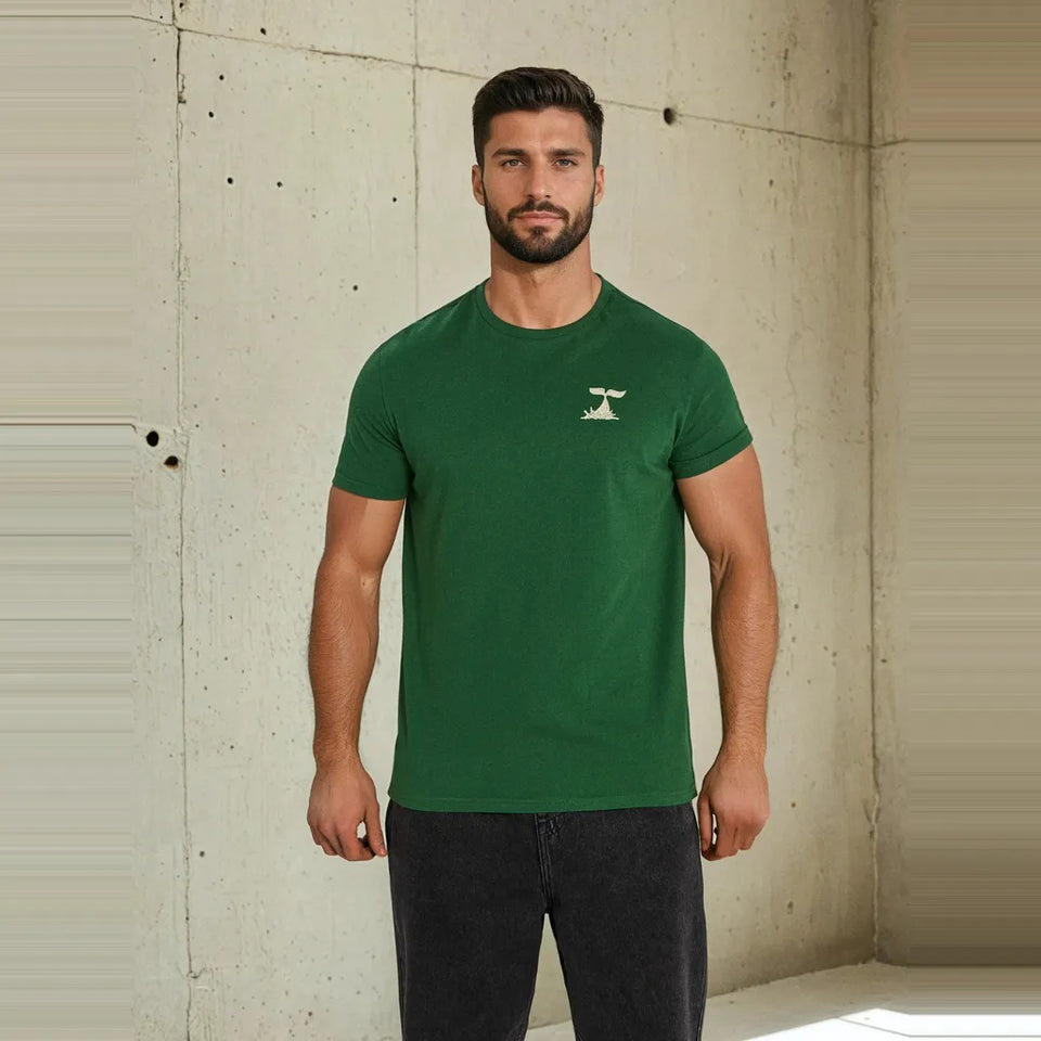 A man wearing a dark green crewneck t-shirt with a small white chest graphic, standing against a textured concrete wall.