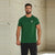 A man wearing a dark green crewneck t-shirt with a small white chest graphic, standing against a textured concrete wall.