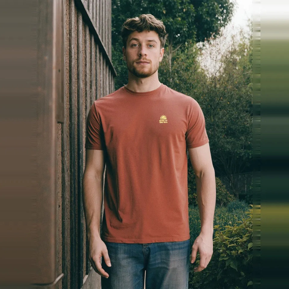 A man wearing a rust-orange crewneck t-shirt with a small yellow logo on the left chest, standing outdoors next to a wooden fence.