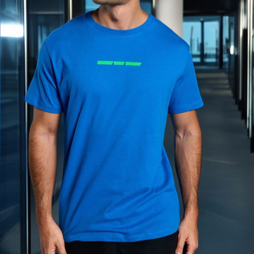 A man wearing a bright blue crewneck t-shirt with a small green graphic text print on the chest, standing in a modern corridor.