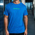 A man wearing a bright blue crewneck t-shirt with a small green graphic text print on the chest, standing in a modern corridor.