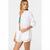 A woman modeling a white long-sleeve button-up shirt with a chest pocket, worn open over a turquoise bikini top and cream shorts, against a plain light gray studio background.