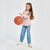 A young girl with long light brown hair stands on a light gray studio background, wearing a light pink long-sleeve ruffled shirt, blue jeans, and white sneakers while holding a basketball.