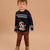 A toddler standing against a solid beige background, wearing a navy blue knit sweater with a white Nordic pattern and a Mickey Mouse graphic in a Santa hat, paired with brown pants and tan boots.
