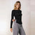 A woman modeling a black long-sleeve off-the-shoulder top with ruching at the side, paired with gray pinstripe trousers, against a light neutral studio background.