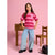 A young girl stands in a studio setting wearing a pink and magenta striped t-shirt with a small white heart graphic on the chest, paired with light-wash wide-leg jeans and black shoes. She is posed in front of a pink background with a wooden table and chair.