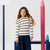A young girl with long wavy brown hair stands in a studio setting wearing a cream long-sleeve top with black horizontal stripes and ruffle details on the shoulders, paired with dark navy wide-leg pants. She is framed by large, stylized pink and blue pencil props against a clean white background.