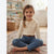 A young girl with a braided ponytail sits cross-legged on a patterned rug in a child's room, wearing a cream knit long-sleeve top with a Peter Pan collar, blue jeans, and a brown belt.