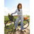 A woman with curly hair stands beside a dark bicycle outdoors on a dirt path, wearing a light blue denim shirt-jacket and matching wide-leg jeans, with a wicker basket on the rear of the bike and a small plant in the front basket.