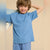 A smiling young boy wearing a light blue short-sleeve crewneck t-shirt and matching blue pants, posing against a warm beige background.