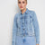A woman modeling a light blue cropped denim jacket with matching light blue denim skirt against a plain light gray studio background.